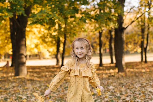 Portrait of a beautiful little blonde girl in a yellow dress in an autumn park holding a bouquet of maple leaves