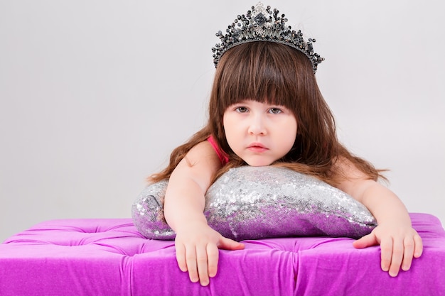 Portrait of beautiful little brunette girl in pink Princess dress with a crown on silver pillow on a gray background. cute baby