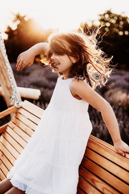 Portrait of a beautiful little girl dancing in the wind against sunset smiling dressed in white dress sitting on a wooden swing in a lavender field.