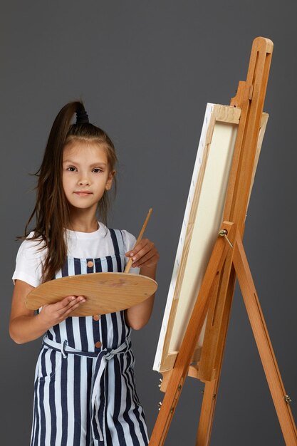 스튜디오 배경에 나무 미술 팔레트와 붓을 들고 있는 아름다운 어린 소녀의 초상화