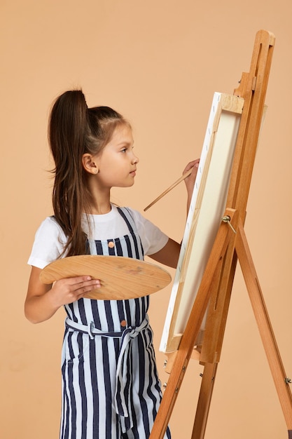 Photo portrait of beautiful little girl holding a wooden art palette and brush on studio background