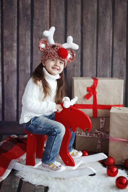 Portrait of a beautiful little girl in the red sweater. Portrait of a cute little girl among the Christmas decorations. Girl rides a reindeer-rocking a hat with horns.