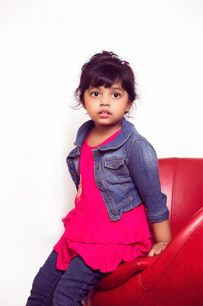 Портрет красивой маленькой девочки, сидящей на красном диване с невинным лицом