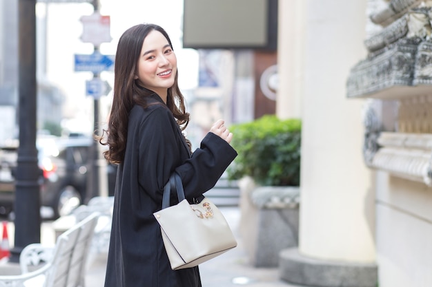 Portrait of a beautiful longhaired Asian female in a black coat carrying a walking bag smiling