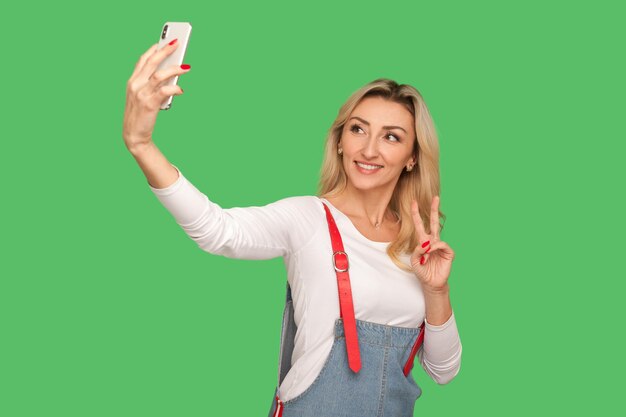 Портрет красивой милой взрослой женщины с дружелюбной улыбкой, делающей селфи на смартфоне и показывающей победный жест в потоковом режиме или в чате в студии видеозвонка, снятой на зеленом фоне