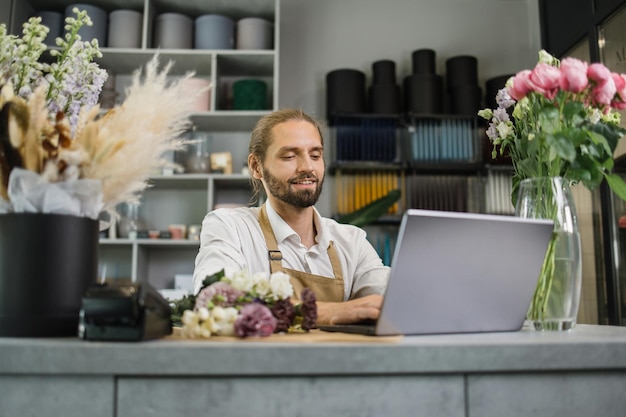 Portrait of beautiful male florist in apron working on laptop in flower store