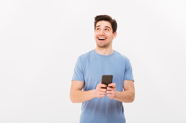 Portrait of beautiful man with short dark hair looking aside and smiling with black cell phone in hand, isolated over white wall