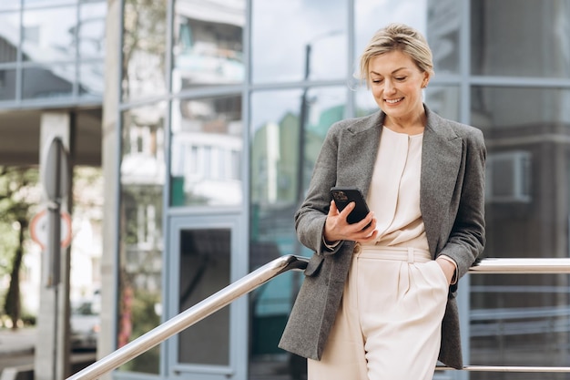 Portrait of a beautiful mature business woman in suit and gray jacket smiling and talking on a phone on the modern urban and office buildings background