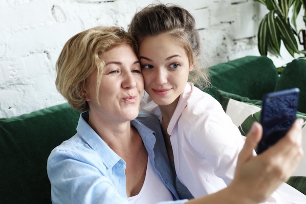 スマートフォンを使用して自分撮りをし、家に笑顔で幸せな美しい成熟した母と娘の肖像画