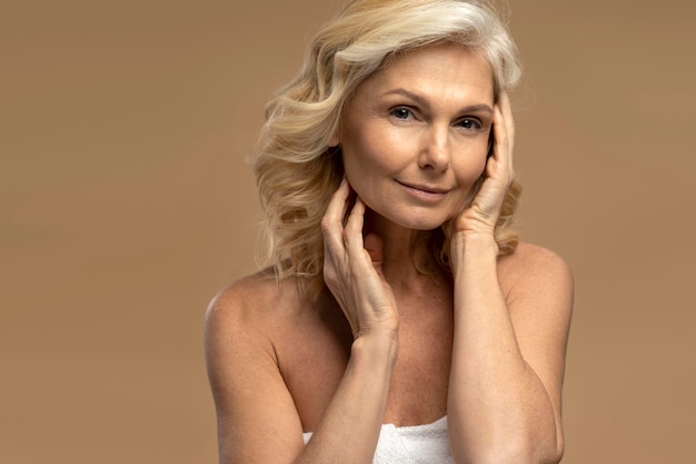 Portrait of beautiful mature woman touching her face looking at mirror isolated on beige background