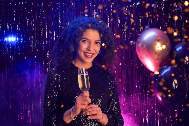 Portrait of beautiful mixed-race woman holding champagne glass and smiling at camera while enjoying party in nightclub, copy space