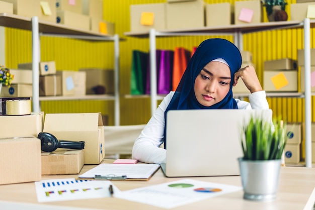 Portrait of beautiful muslim owner asian woman freelancer sme business online shopping working on laptop computer with parcel box on table at home - Business online shipping and delivery