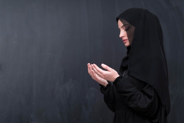 Portrait of beautiful muslim woman in fashionable dress with hijab making traditional prayer to God keeps hands in praying gesture isolated on black background. High quality photo