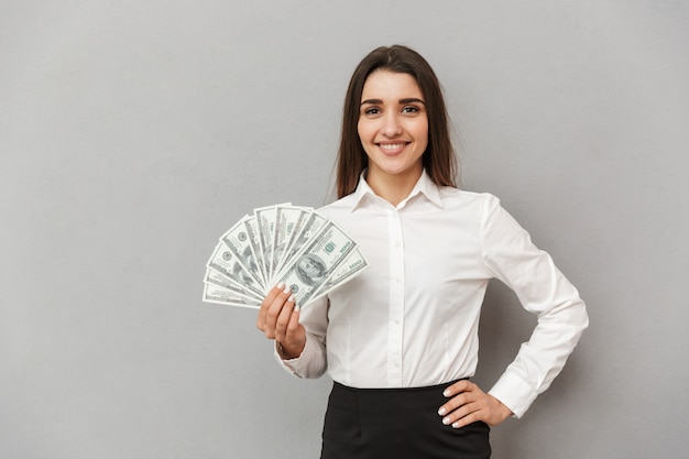 Portrait of beautiful office woman with long brown hair in business wear smiling and holding fan of money, isolated over gray wall