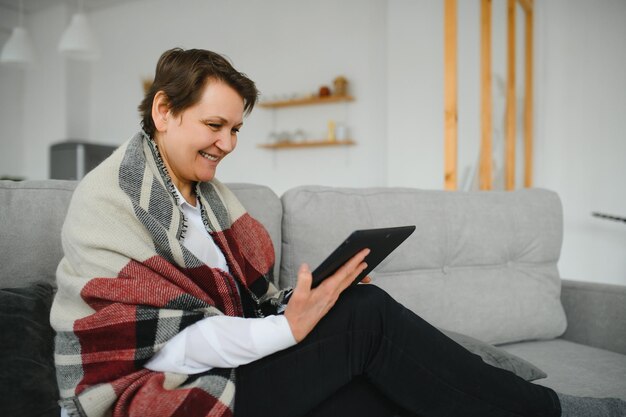 Portrait of beautiful old woman using tablet pc and smiling sitting on sofa at home