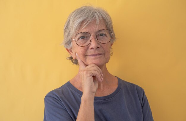 Portrait of beautiful pensive senior woman in black looking at camera wearing eyeglasses isolated on yellow background