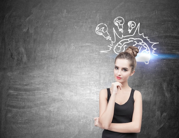 Portrait of a beautiful and pensive young woman wearing a black tank top. Her hair is in a bun. She is thinking and half smiling. Blackboard background with a brain sketch and light bulbs. Mock up