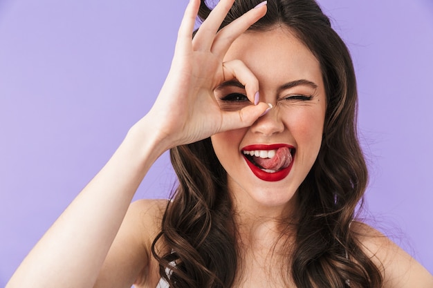 Portrait of a beautiful pin-up girl wearing bright makeup standing isolated over violet wall, showing ok