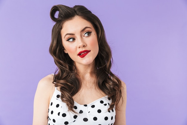 Portrait of a beautiful pin-up girl wearing bright makeup standing isolated over violet wall, thinking