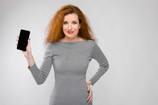 Portrait of beautiful redhead happy smiling young woman in gray clothes showing mobile phone 