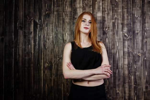 Ritratto di una bella ragazza dai capelli rossi in cima nera e gonna nera in posa in studio accanto alla parete di legno.