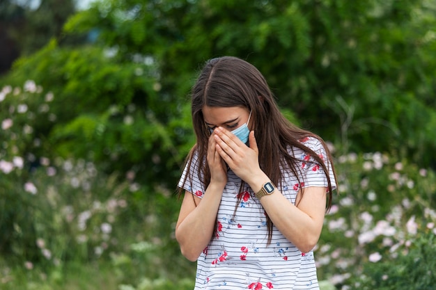 Portrait of beautiful sad woman in medical mask sneezes among white flowers