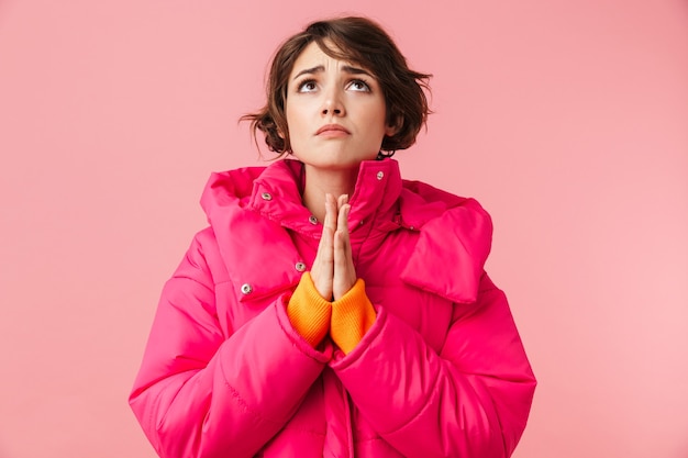 Portrait of beautiful sad woman in warm coat looking upward and holding palms together isolated on pink