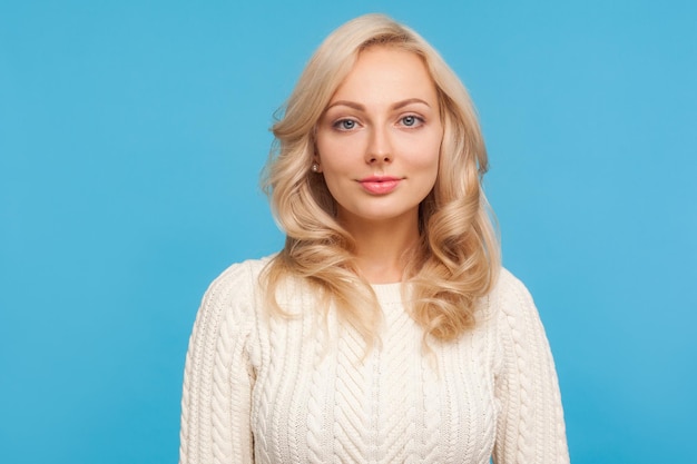 Portrait of beautiful self confident woman with curly blond hair looking at camera with light smile on face. Indoor studio shot isolated on blue background