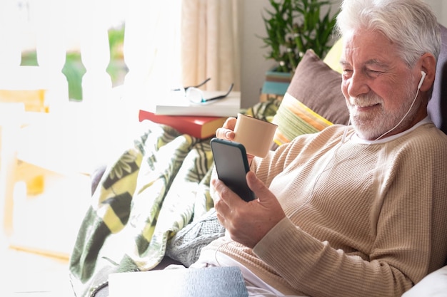 Portrait of beautiful senior man with earphones lying down on sofa at home using phone drinking a coffee cup Bright light from the window Retired seniors using wireless technology