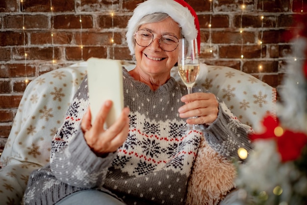 Portrait of beautiful senior woman in santa hat video chatting by mobile phone at Christmas time holding a champagne flut elderly happy lady sitting alone at home using wireless technology