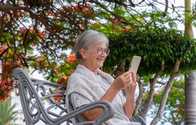 Portrait of beautiful senior woman sitting relaxed in a park\
bench using mobile phone elderly lady enjoing tech and social\
concept of serene retirement