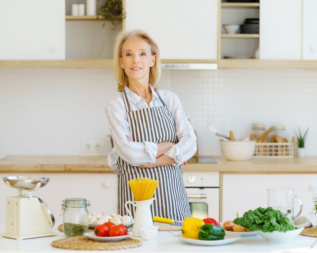 Ritratto di bella donna anziana in piedi in cucina e sorride alla macchina fotografica mentre si prepara