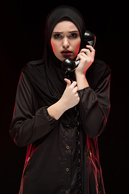 Portrait of beautiful serious scared frightened young muslim woman wearing black hijab calling for help on black