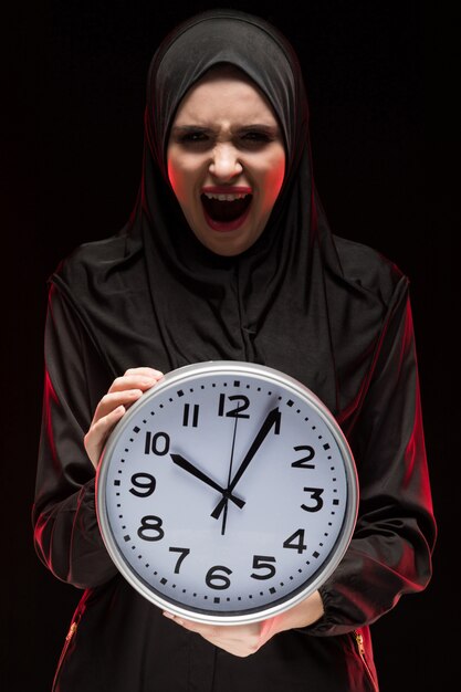 Portrait of beautiful serious scared frightened young muslim woman wearing black hijab holding clock