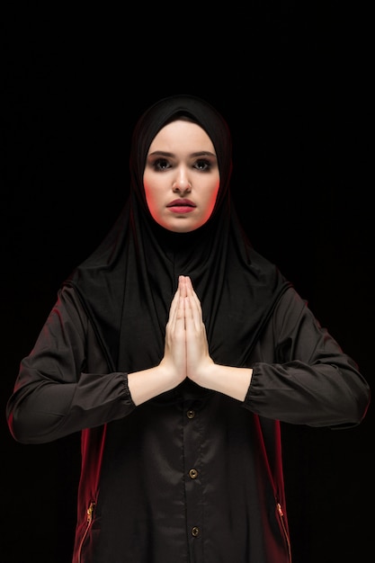 Portrait of beautiful serious young muslim woman wearing black hijab with hands near her face as praying 