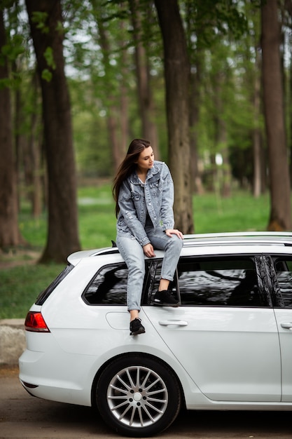 Portrait of beautiful sexy fashion brunette woman model in jeans jacket sitting on luxury white car and relaxes. Fashion concept. Idea for girl photoshoot with car. Vertical.