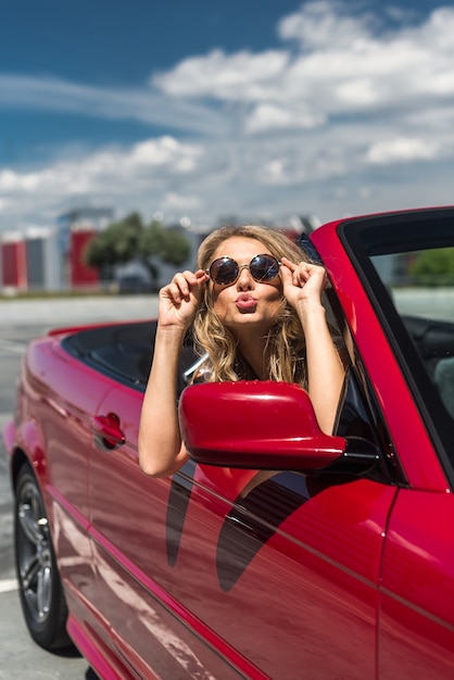 바다와 하늘 배경으로 럭셔리 빨간색 컨버터블 자동차에 앉아 선글라스에 아름 다운 섹시 패션 여자 모델의 초상화. 화창한 여름날에 여행을 운전하는 젊은 여자. 바다와 하늘. 빨간 cabrio.