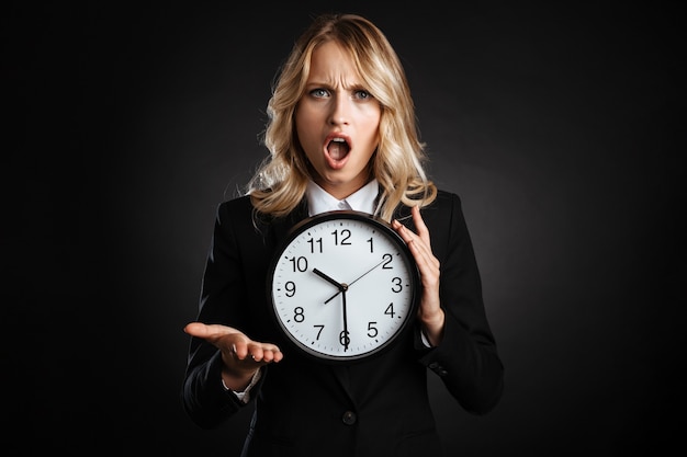 Portrait of a beautiful shocked blonde haired business woman dressed in formal clothes standing isolated over black wall, showing alarm clock