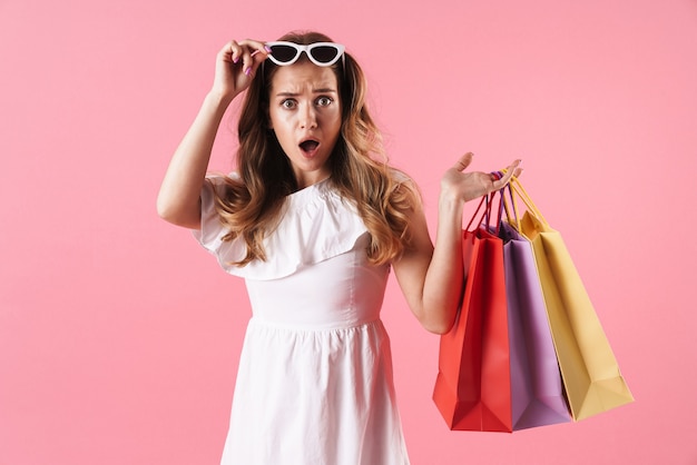 Portrait of beautiful shocked woman wearing white dress holding shopping bags and sunglasses isolated over pink wall