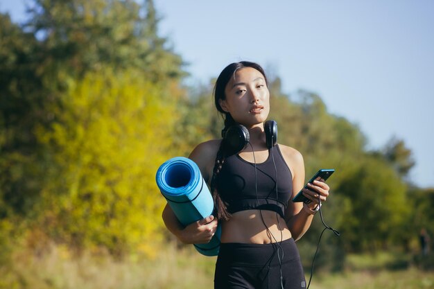 Portrait of a beautiful slender Asian woman, athlete in the woods, listening to music with headphones and holding a yoga mat, athlete smiling and looking at the camera