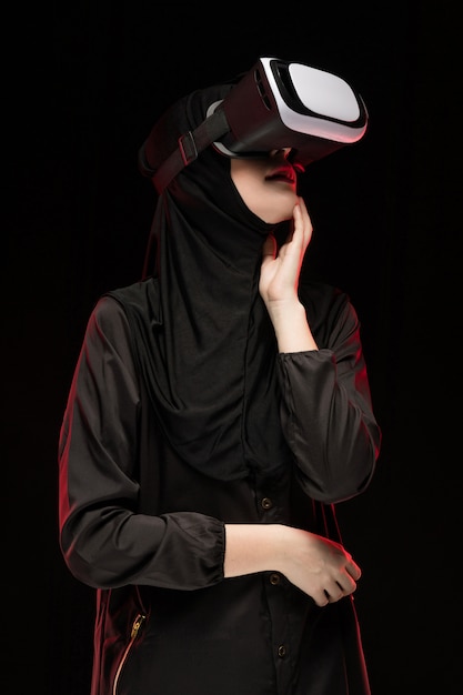 黒の背景に仮想現実のヘッドセットを使用して黒のヒジャーブを着ている美しいスマートな若いイスラム教徒の女性の肖像画