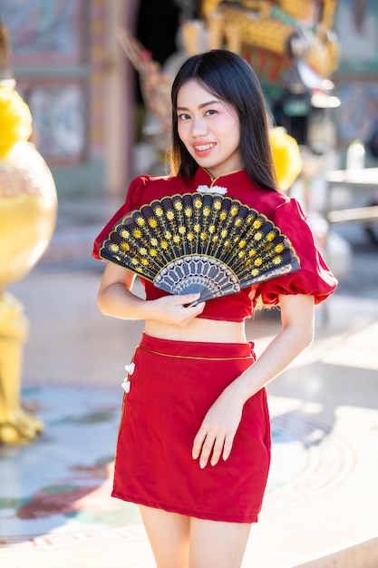 肖像画美しい笑顔赤い伝統的な中国のチャイナドレスの装飾を身に着けて、タイの中国の神社で中国の旧正月祭りのための中国のファニングを開催しているアジアの若い女性