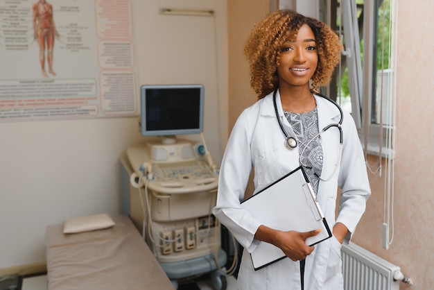Портрет красивой улыбающейся женщины-афро-американского доктора, стоящей в медицинском кабинете
