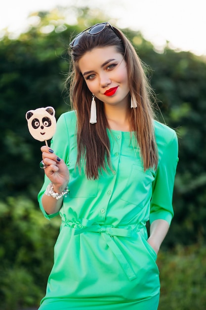 Photo portrait of beautiful smiling girl in green dress, showing at camera candy like panda on stick in hand. stylish woman with red lips and long earrings, holding hand in pocket and posing outdoor.