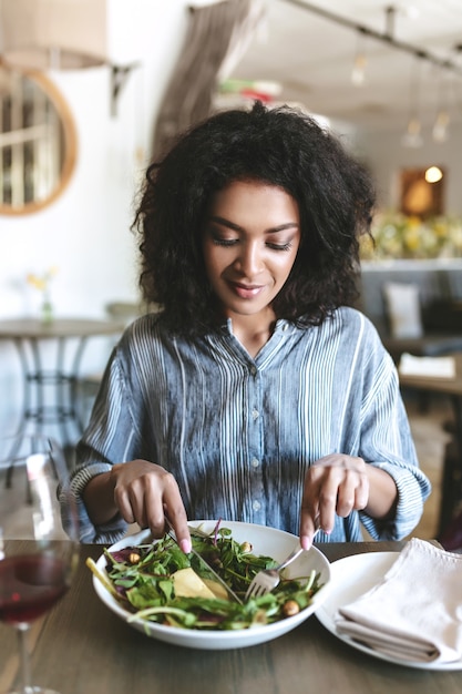 portrait of beautiful smiling girl with dark curly hair sitting in restaurant with glass of red wine and salad on table
