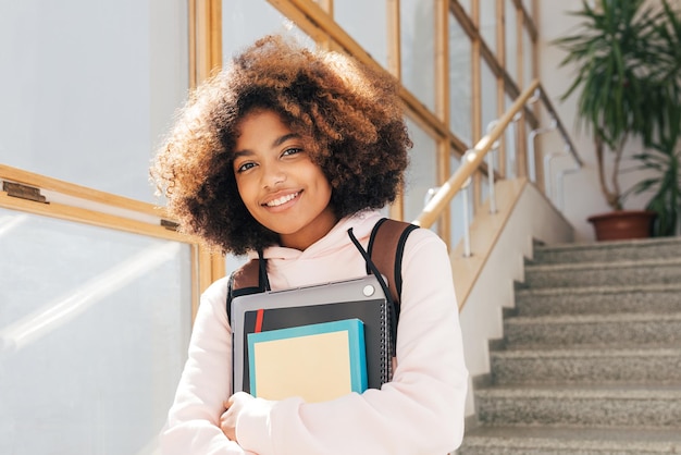 Портрет красивой улыбающейся девушки с ноутбуком и книгами, стоящей в школе на лестнице и смотрящей в камеруx9xA