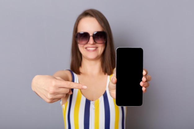 Portrait of beautiful smiling satisfied caucasian woman in bright swimsuit standing isolated over gray background showing smart phone with empty display advertising travel agency