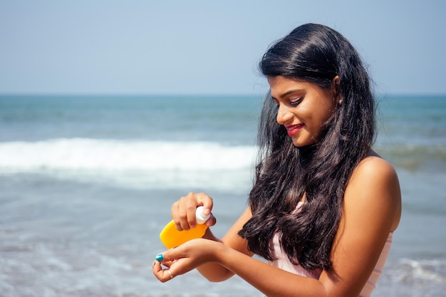 Ritratto di una bella e sorridente sorriso bianco come la neve donna indiana capelli neri e pelle scura in una maglietta rosa con in mano una bottiglia di crema solare spray sulla spiaggia che si gode la vacanza del paradiso del corpo spf