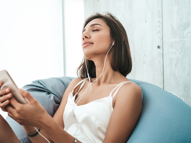 Portrait of beautiful smiling woman dressed in white pajamas and listening to music with headphones. Carefree model sitting on soft bag chair. 