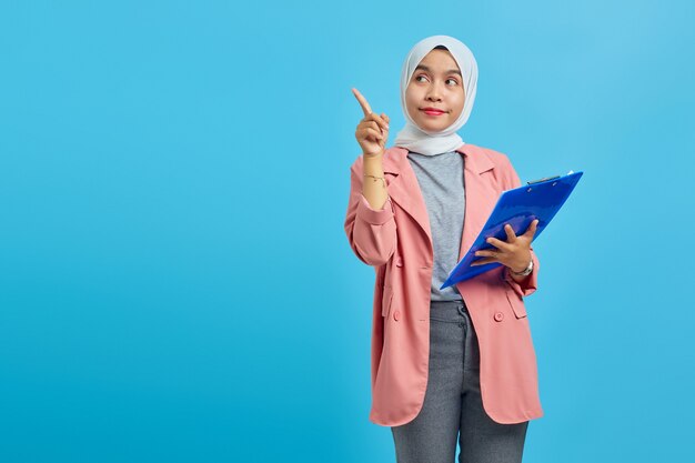 Portrait of a beautiful smiling woman holding a folder standing\
and pointing to an empty space on a blue background